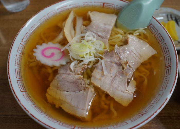 An overhead image of a bowl of ramen with a clear light brown broth, several slices of meat, and a white fishcake with a pink swirl